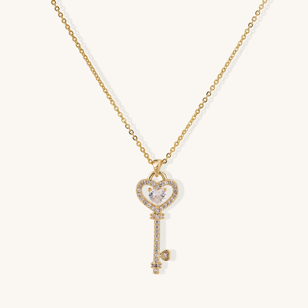 Gold-plated key pendant necklace with heart top and crystal embellishments on adjustable chain against cream background