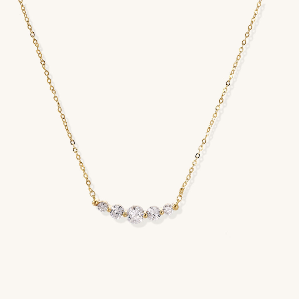 Delicate 14K gold plated necklace with curved arc of sparkling zirconia stones on cream background.