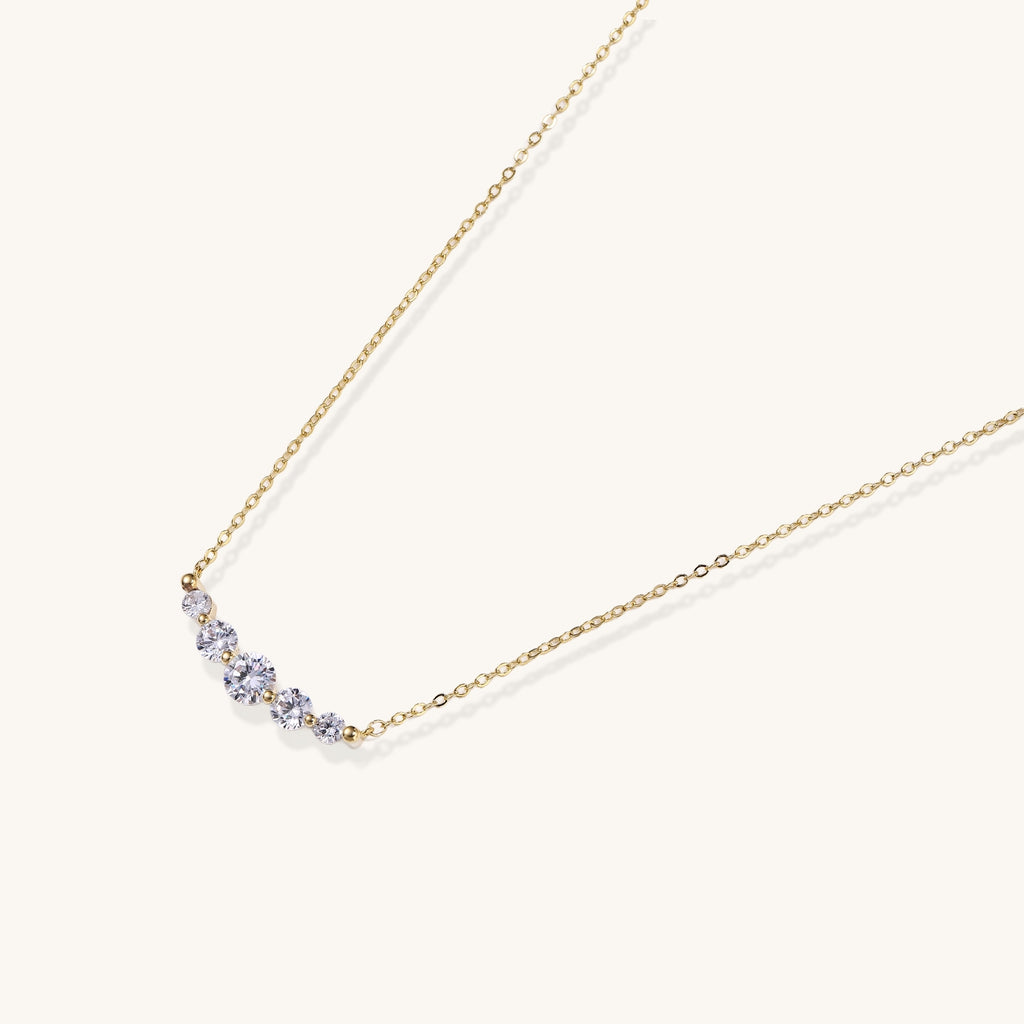 Delicate 14K gold plated necklace with curved arc of sparkling zirconia stones on cream background.