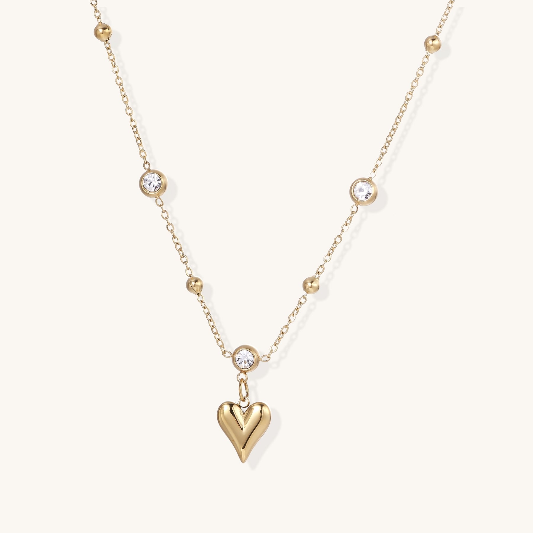 Dainty 14K gold plated necklace with heart pendant and bezel-set crystals on delicate chain against cream background.